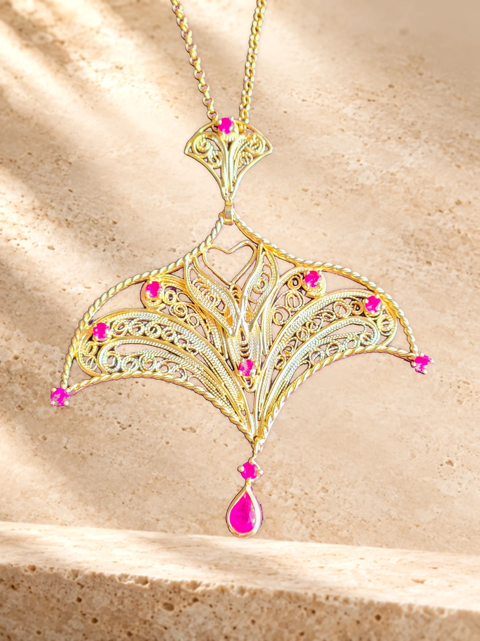 Source-preserving editorial composite of the MELİHA necklace on a warm stone background: curved filigree motifs, pink-red accents and a teardrop-shaped pendant.